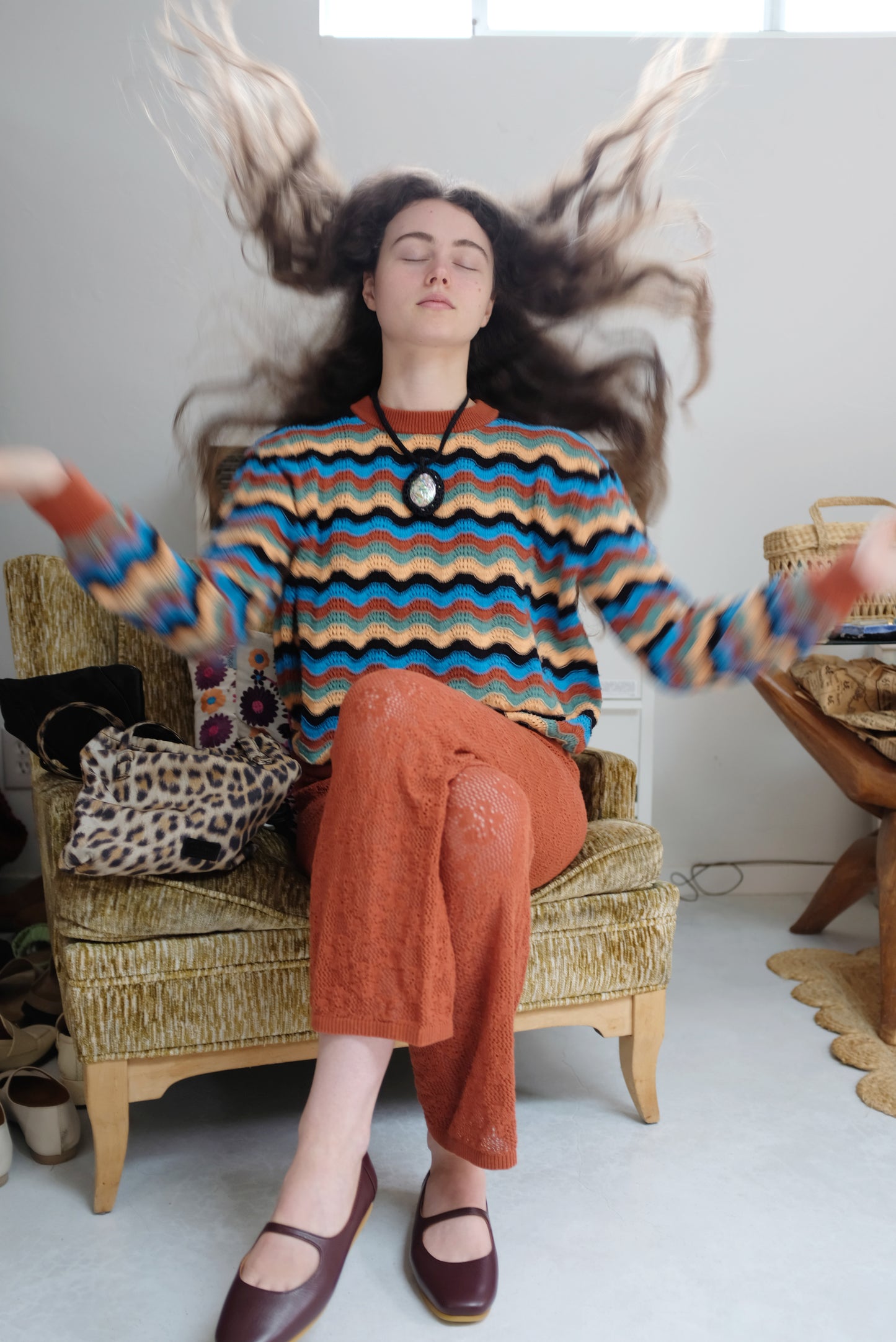 Person wearing a colorful striped sweater and orange pants sitting on a chair with long hair flowing.